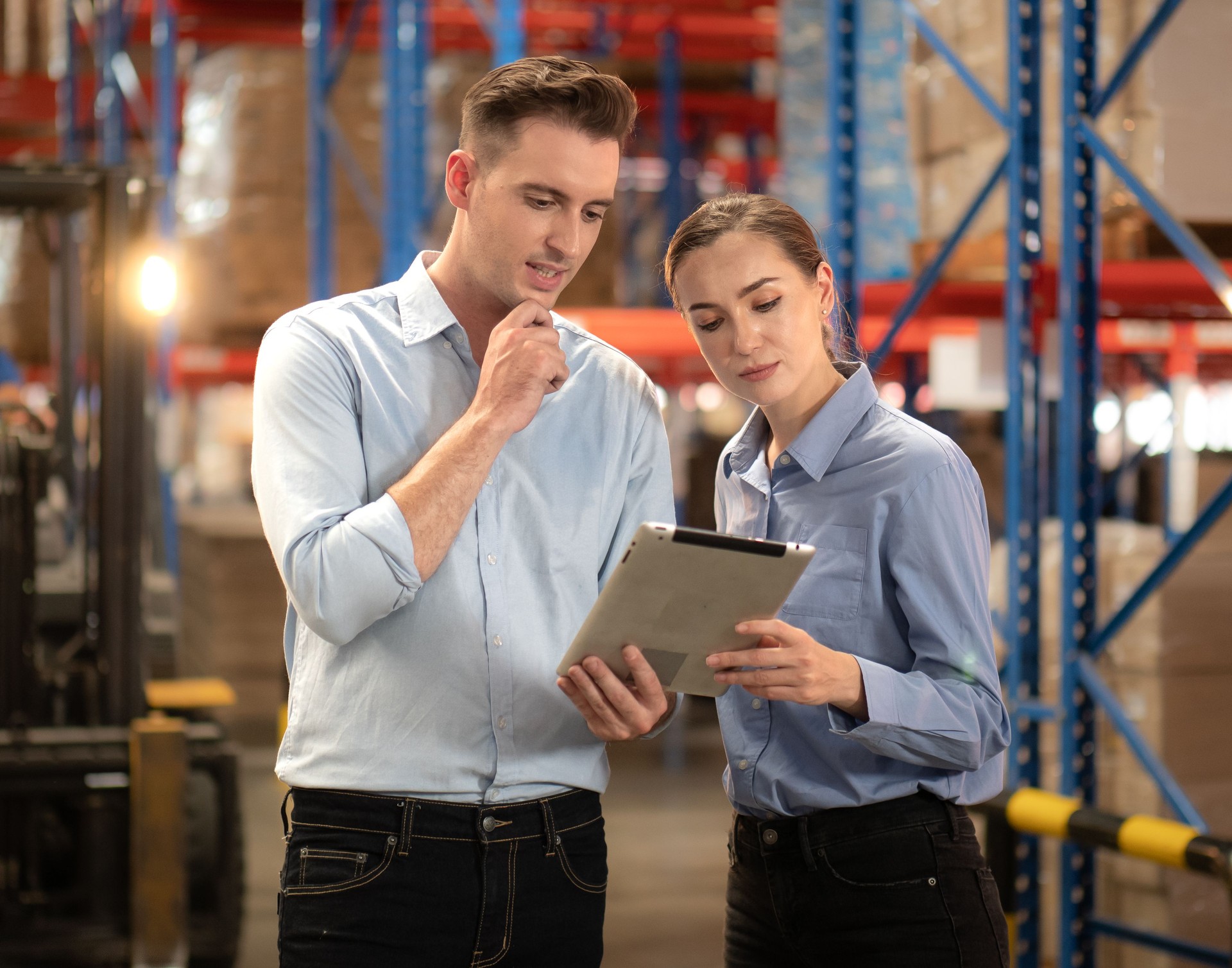 Distribution warehouse manager and client businesswoman using digital tablet checking inventory storage on shelf.